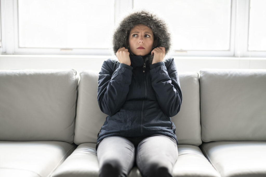 Woman sitting on gray couch, wearing black winter coat with furry hood pulled up.